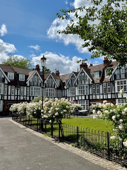 A vibrant urban garden featuring a dense bed of blooming pink and peach roses with lush green foliage in the foreground. Behind the roses are various trees with autumn-colored leaves and well-maintain